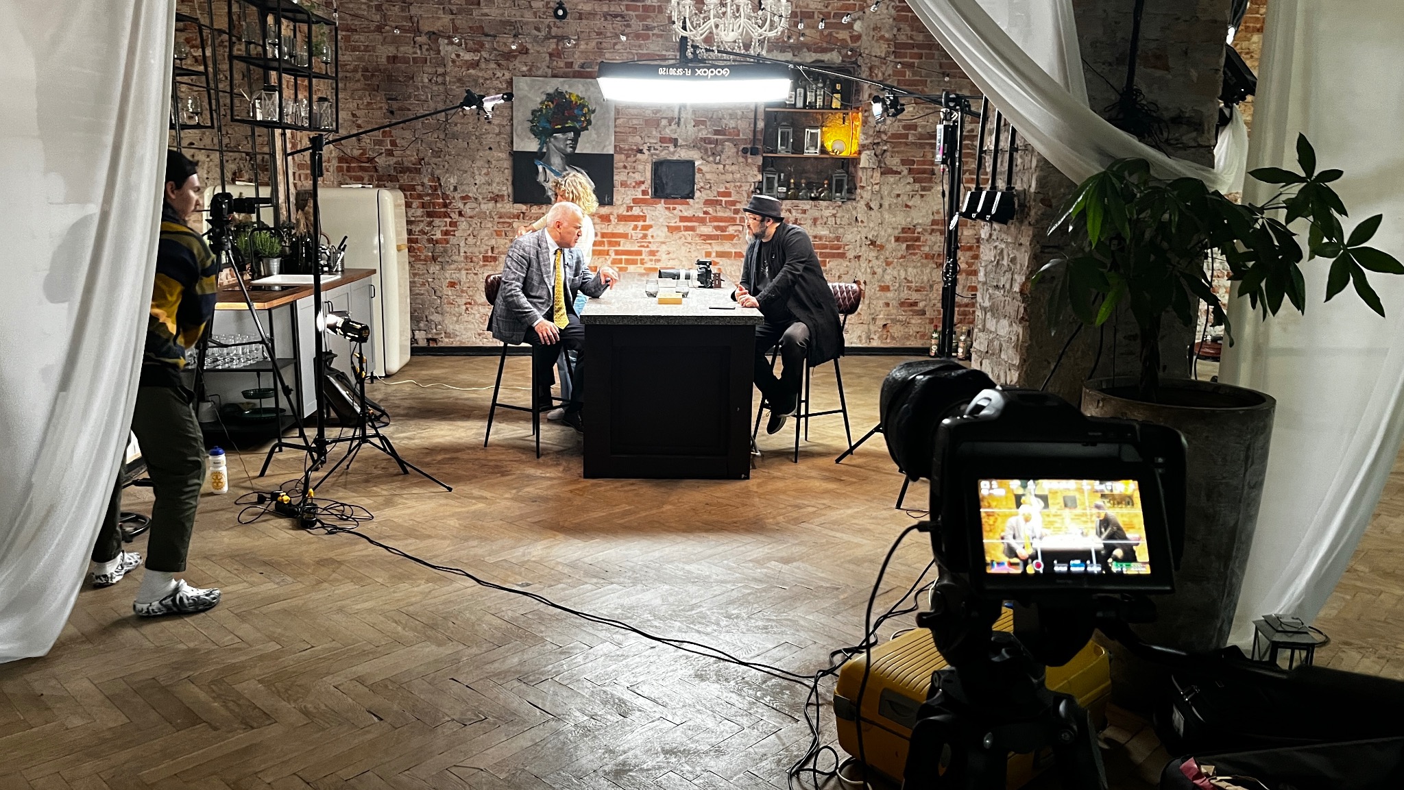 Plan filmowy w loftowym wnętrzu z ceglaną ścianą. Dwóch mężczyzn siedzi przy ciemnym stole oświetlonym lampą studyjną. W tle regał z dekoracjami i biała lodówka. Ujęcie z perspektywy kamery...