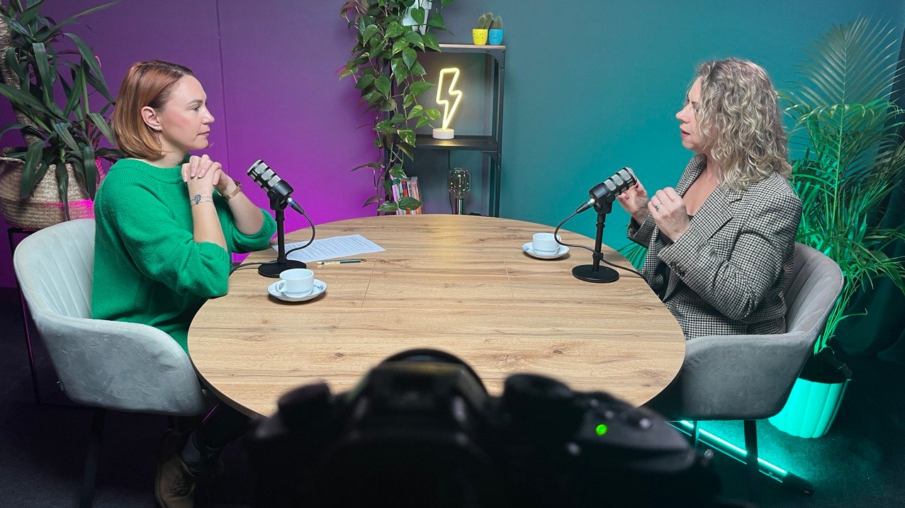 Dwie kobiety prowadzą rozmowę przy okrągłym stole z mikrofonami, filiżankami kawy i dokumentami, w tle rośliny i neon w kształcie błyskawicy, widok zza kamery.