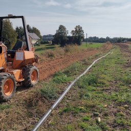 Amplus Arkadiusz Mazurek Wykopy koparkami Alarmy Elektryka Monitoring - Pomarańczowy traktor rowokoparka w trakcie pracy na polu, układający rurę. Widoczne świeżo wykopane bruzdy w ziemi. W tle zabudowania wiejskie.