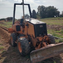 Amplus Arkadiusz Mazurek Wykopy koparkami Alarmy Elektryka Monitoring - Pomarańczowy równiarka w trakcie wykonywania wykopu w polu, widoczna długa bruzda. Maszyna z przodu, w tle zabudowania i drzewa, pochmurne niebo.