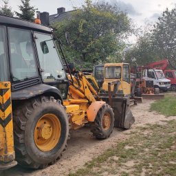 Amplus Arkadiusz Mazurek Wykopy koparkami Alarmy Elektryka Monitoring - Żółto-czarna koparko-ładowarka Volvo na żwirowym placu, z rzędem maszyn budowlanych w tle, w otoczeniu zieleni i drzew, pochmurne niebo.