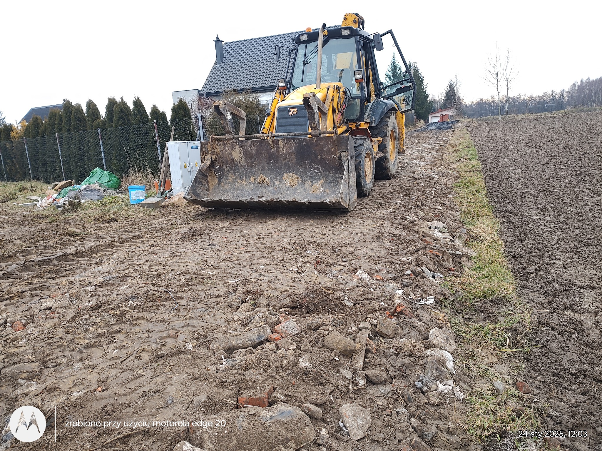 Żółta koparko-ładowarka JCB na nierównym, błotnistym terenie budowy z gruzem, dom w tle. Widoczne ślady opon i świeżo przekopana ziemia. Krajobraz po niwelacji.