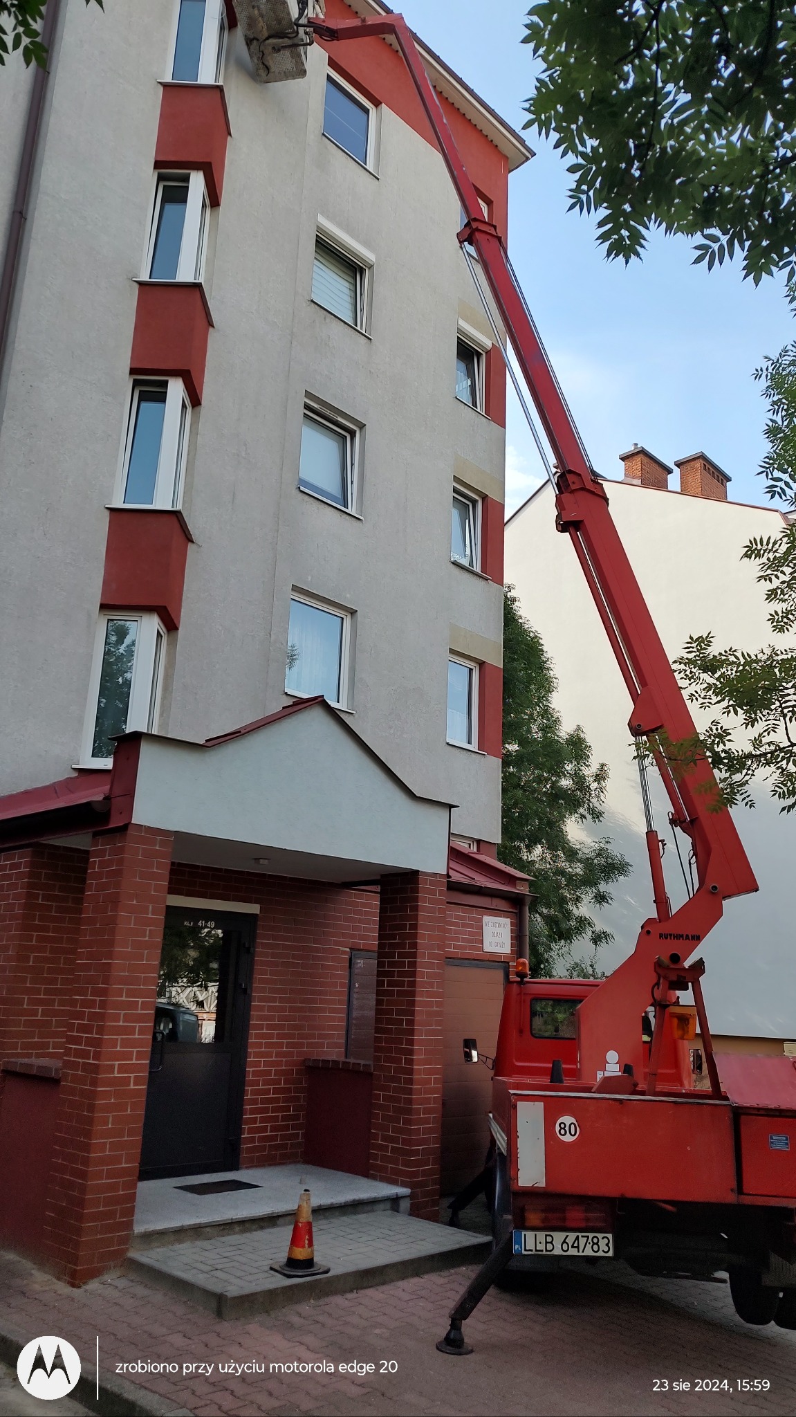 Czerwony podnośnik koszowy Ruthmann przed budynkiem mieszkalnym, potencjalnie używany do prac na wysokości, z widocznym stożkiem ostrzegawczym.