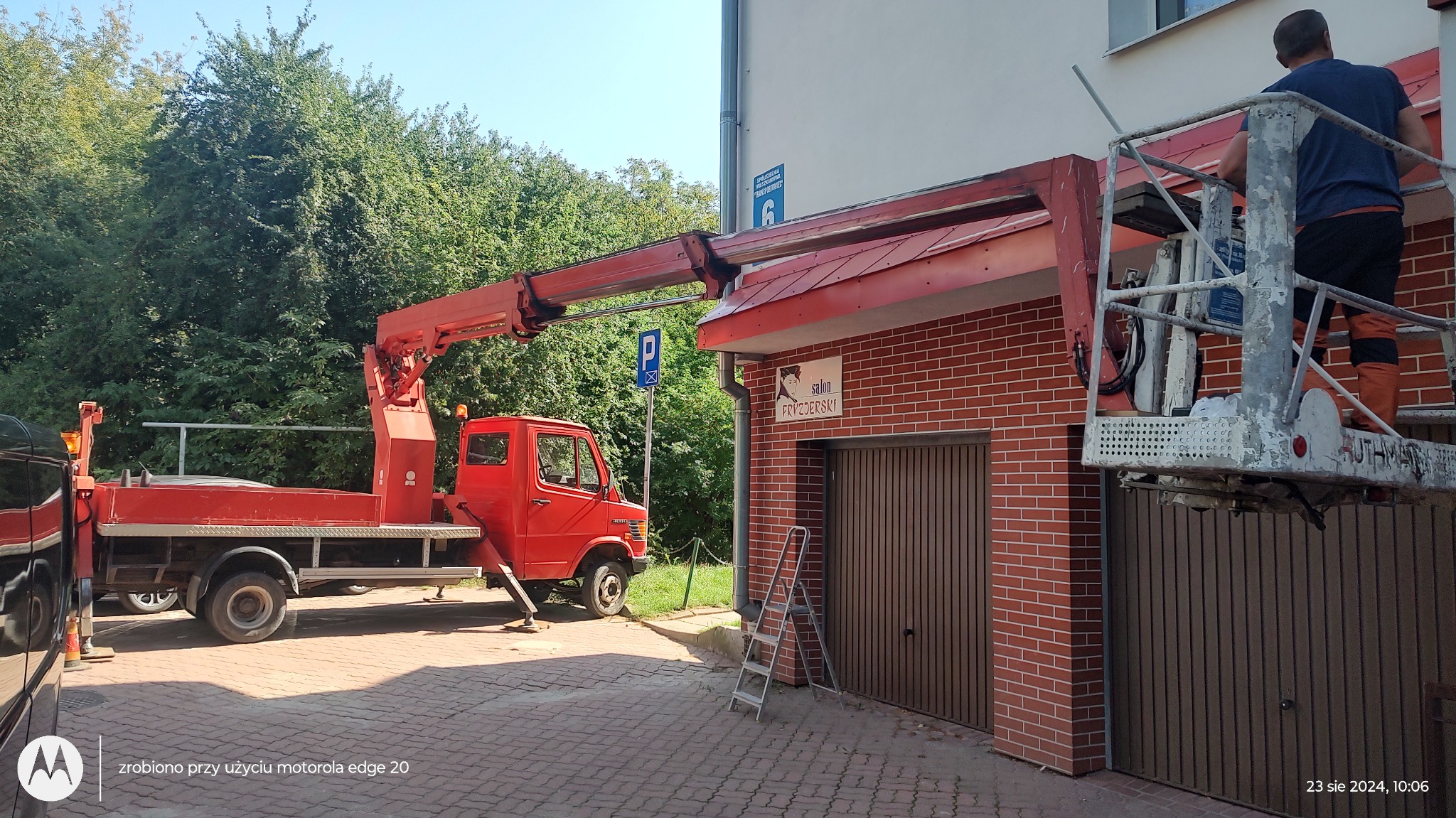 Prace na wysokości z użyciem podnośnika koszowego przy budynku salonu fryzjerskiego; widoczny czerwony samochód z podnośnikiem i pracownik na platformie.