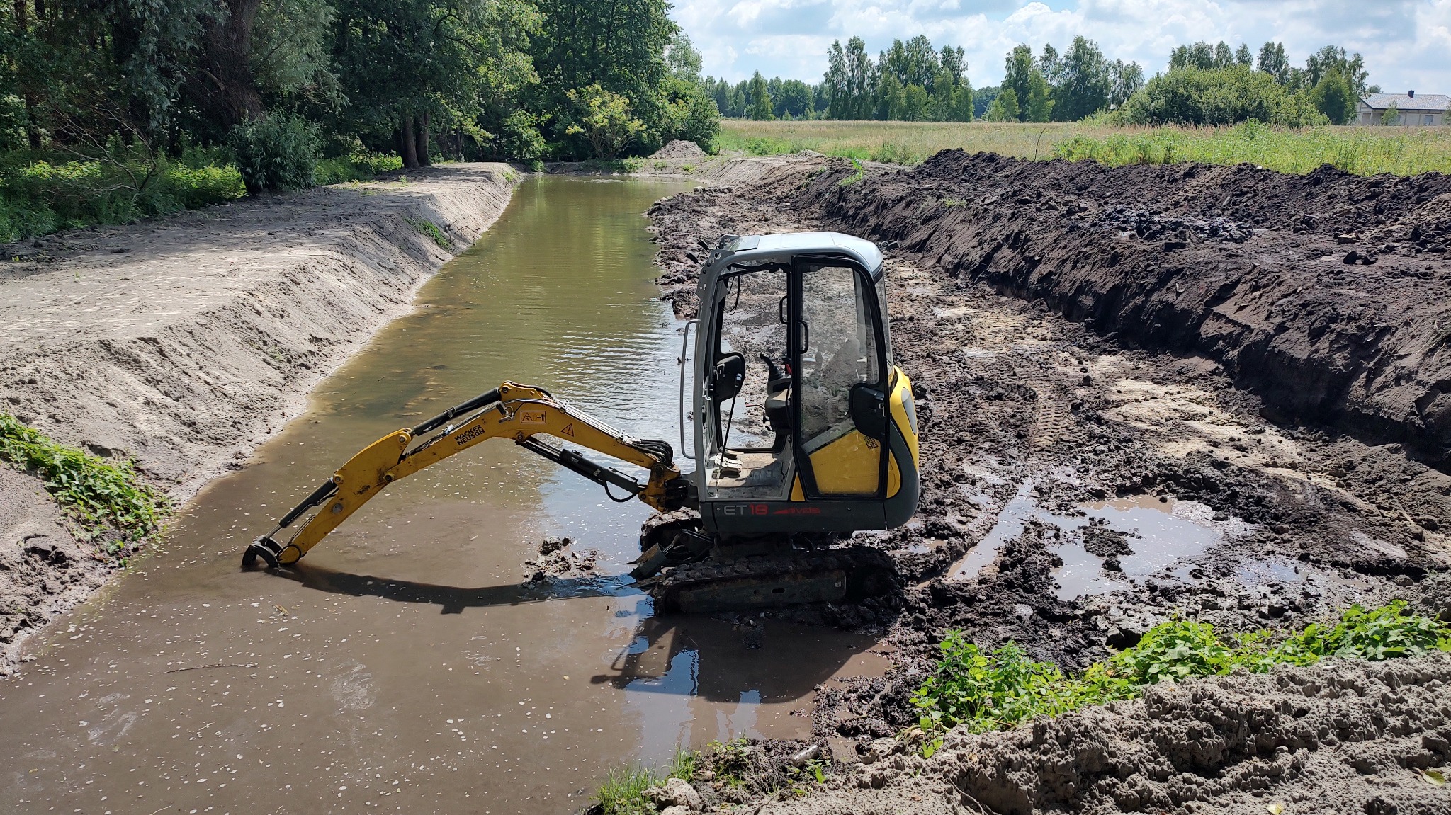 Żółta minikoparka w trakcie prac ziemnych przy rowie melioracyjnym, widoczne ślady gąsienic i świeżo wykopana ziemia. W tle zieleń drzew i łąka.
