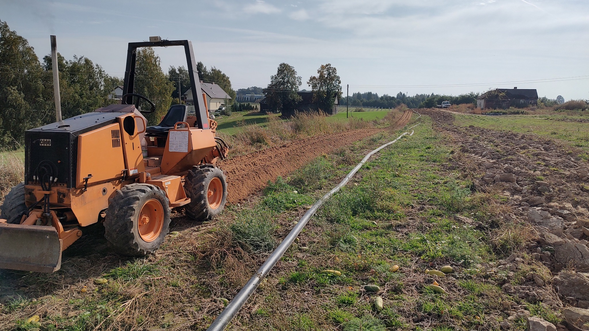 Pomarańczowy traktor rowokoparka w trakcie pracy na polu, układający rurę. Widoczne świeżo wykopane bruzdy w ziemi. W tle zabudowania wiejskie.
