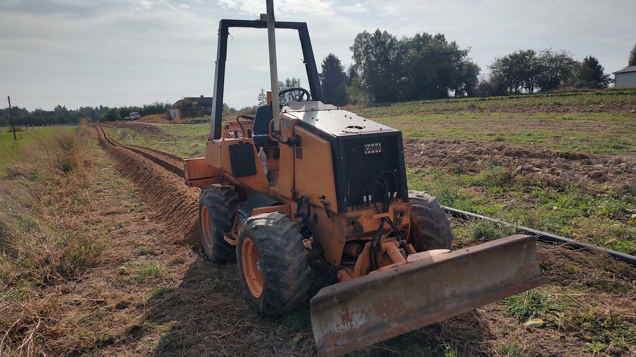 Pomarańczowy równiarka w trakcie wykonywania wykopu w polu, widoczna długa bruzda. Maszyna z przodu, w tle zabudowania i drzewa, pochmurne niebo.