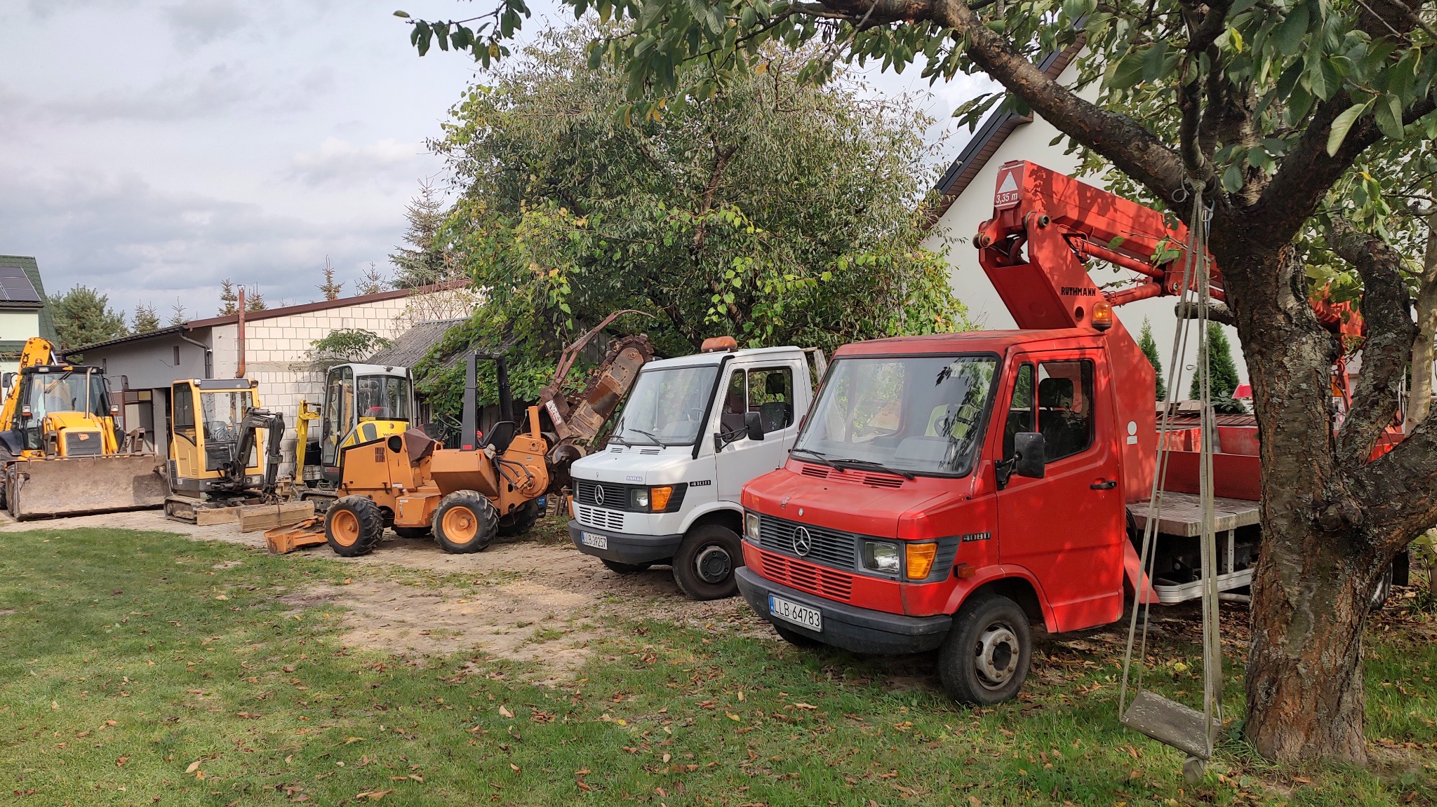 Różnorodny sprzęt budowlany: koparki, minikoparki i podnośniki koszowe Mercedes-Benz, ustawione na trawiastym terenie, w tle budynki i drzewa.