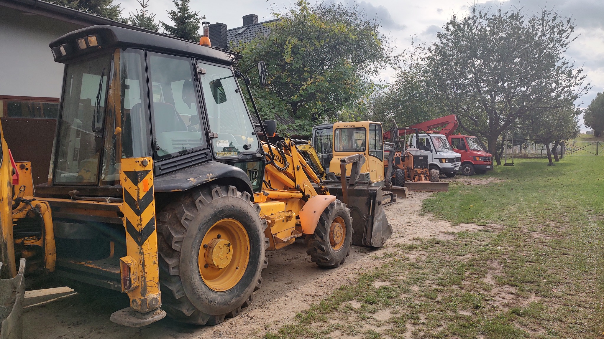 Żółto-czarna koparko-ładowarka Volvo na żwirowym placu, z rzędem maszyn budowlanych w tle, w otoczeniu zieleni i drzew, pochmurne niebo.