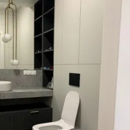 Nowoczesna łazienka w odcieniach szarości i bieli z wiszącą toaletą, bidetem i szafkami w zabudowie, oświetlona lampą z trzema kulami.