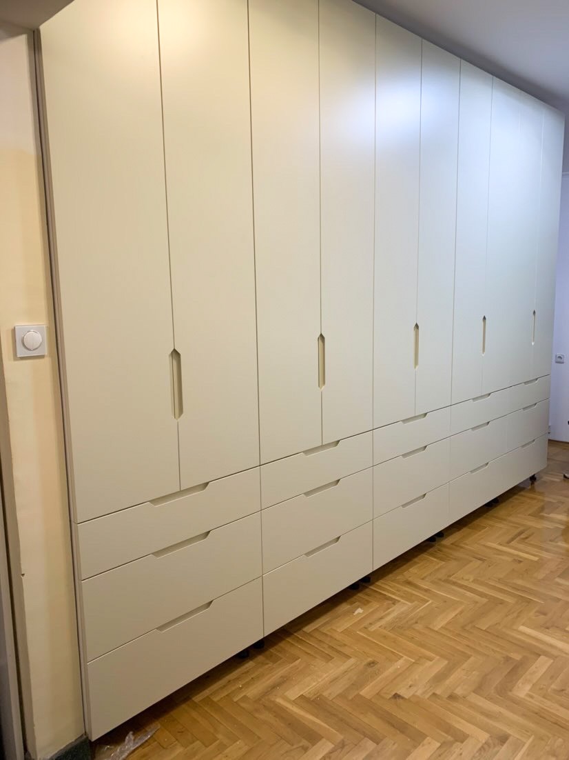 Duża, kremowa szafa wnękowa z minimalistycznymi uchwytami, obejmująca całą ścianę, z dolnymi szufladami i parkietem w jodełkę na podłodze.