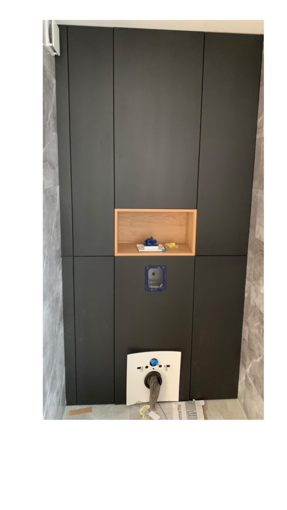 Ciemna zabudowa meblowa w łazience, z otwartą wnęką i przygotowanym miejscem na montaż toalety podwieszanej, widoczne elementy instalacji.