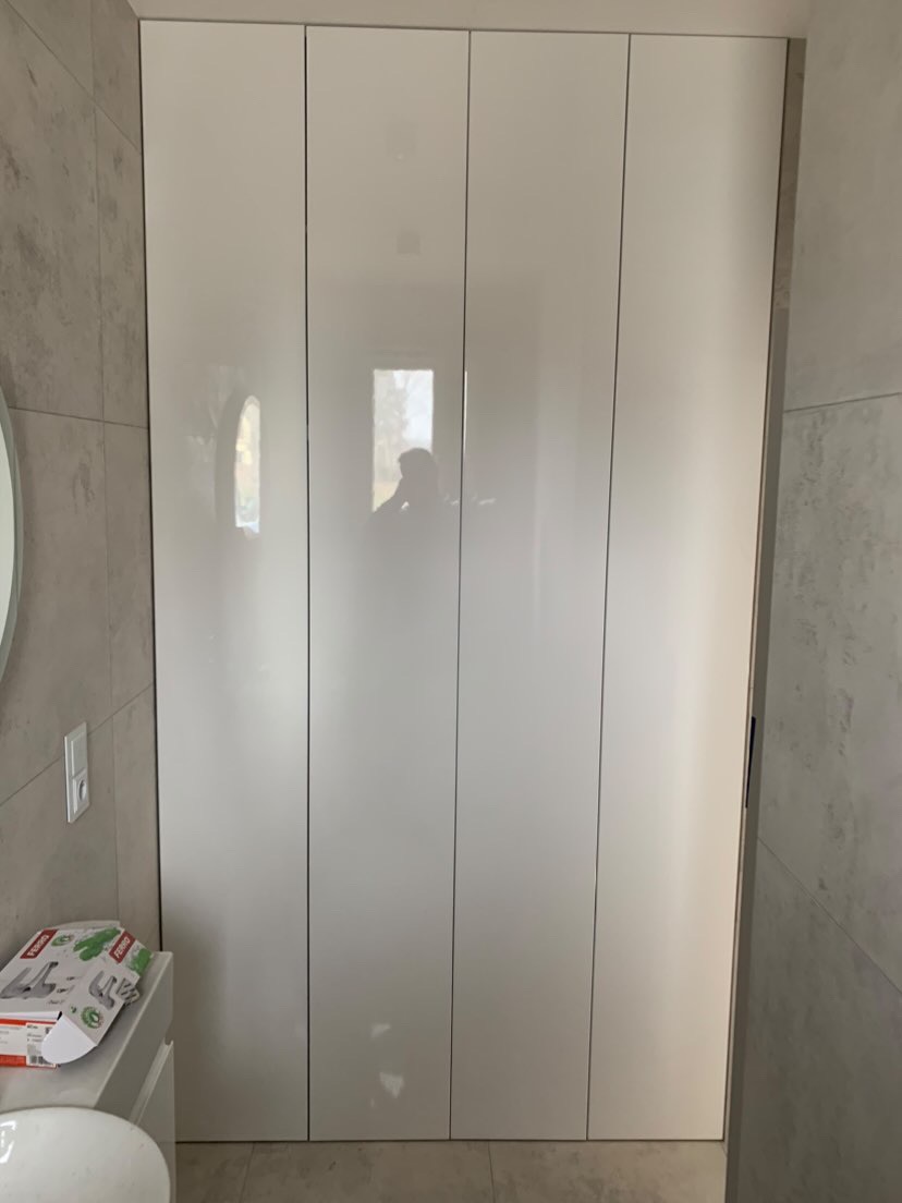 Biała, wysoka szafa wnękowa z czterema frontami na wysoki połysk, w łazience wykończonej płytkami imitującymi beton, z widocznym odbiciem osoby w drzwiach.