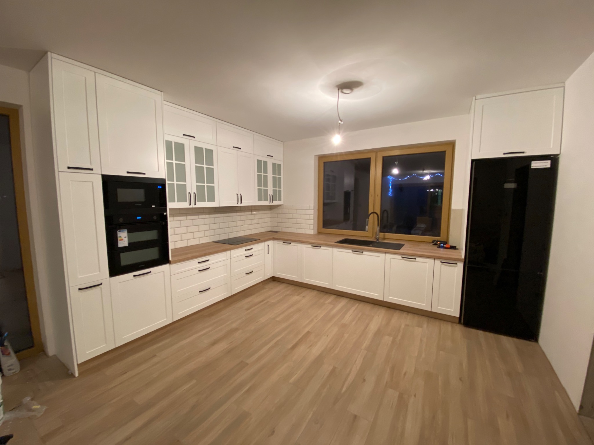 Nowoczesna kuchnia z białymi frontami, drewnianym blatem i czarnymi akcentami, w tym piekarnik i lodówka, oświetlona pojedynczą lampą wiszącą.