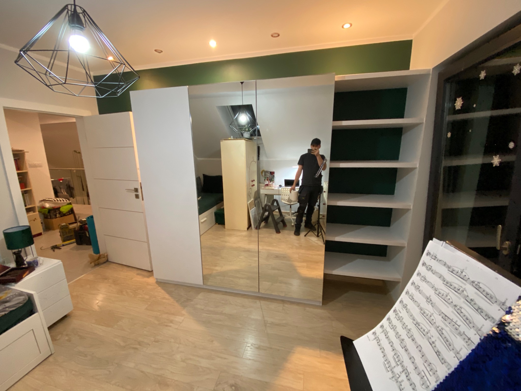 Biała szafa na wymiar z lustrzanymi drzwiami przesuwnymi, w tle widoczna osoba robiąca zdjęcie, w pomieszczeniu lampa wisząca o geometrycznym kształcie.