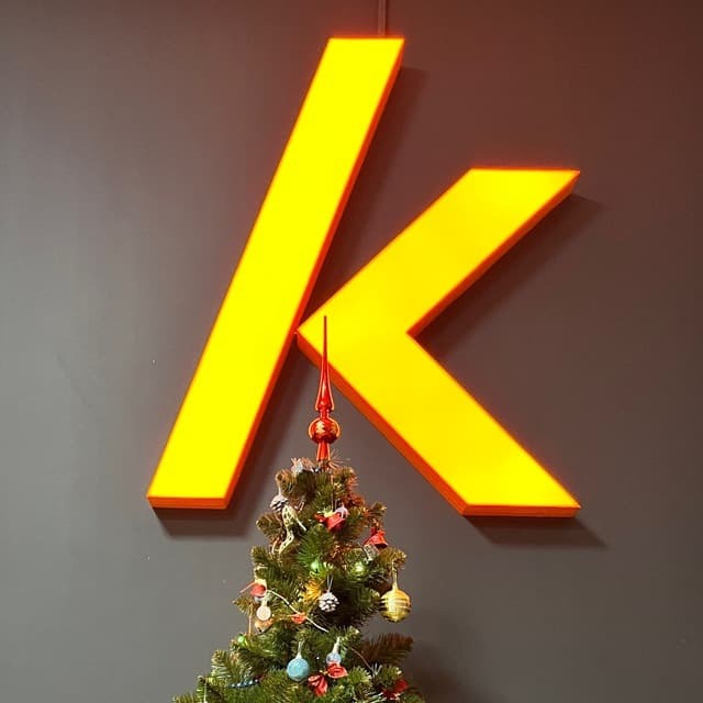 Żółty, podświetlany znak 'K' na szarej ścianie, z choinką w dolnej części kadru. Dekoracje świąteczne w biurze.
