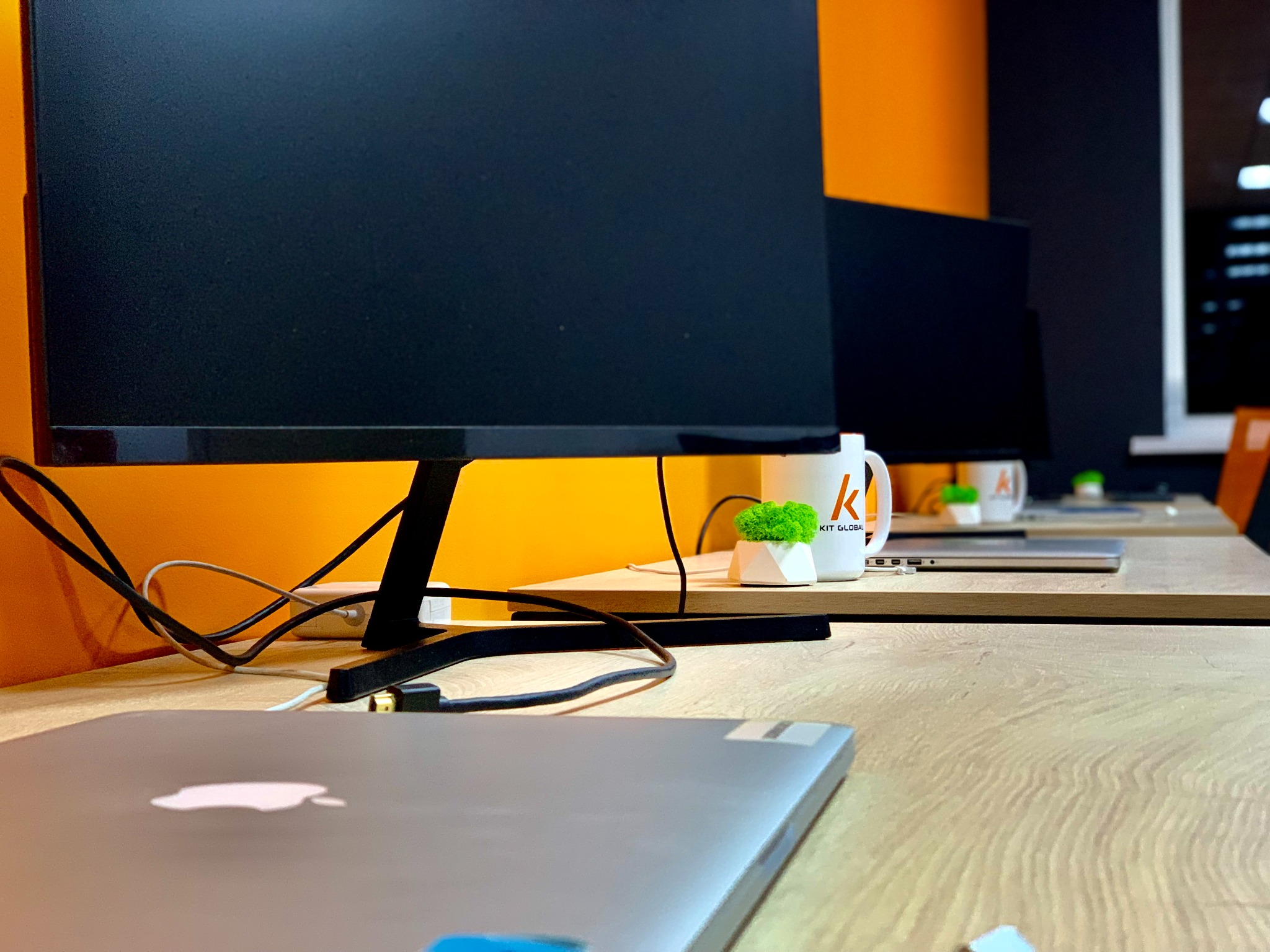 Stanowisko pracy z komputerami, kubkami z logo firmy Kit Global i dekoracyjnymi roślinkami na jasnym biurku, pomarańczowa ściana w tle.
