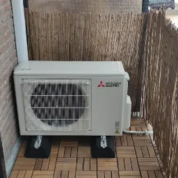 Zewnętrzna jednostka klimatyzacji Mitsubishi Electric zamontowana na balkonie z drewnianą podłogą i bambusową osłoną. Widoczne czarne podkładki antywibracyjne pod jednostką.