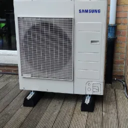 Zewnętrzna jednostka klimatyzacji Samsung na drewnianym tarasie, zamontowana na czarnych gumowych podstawach, z widoczną rurą spustową i ceglaną ścianą budynku w tle.