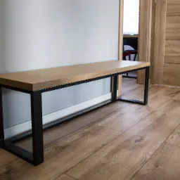 Prosta ławka z drewnianym blatem i czarną metalową podstawą, ustawiona w przedpokoju na drewnianej podłodze, obok drzwi z tego samego drewna.