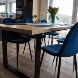 Stół z blatem drewnianym o jasnym odcieniu i czarnymi metalowymi nogami, otoczony czterema granatowymi krzesłami z weluru, na stole okrągłe podkładki i wazon z tulipanami, w tle nowoczesna kuchnia...