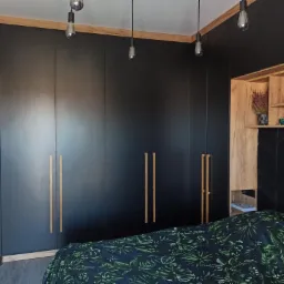 Czarna zabudowa szafy z drewnianymi uchwytami i oświetleniem w sypialni, widoczna pościel w liście.