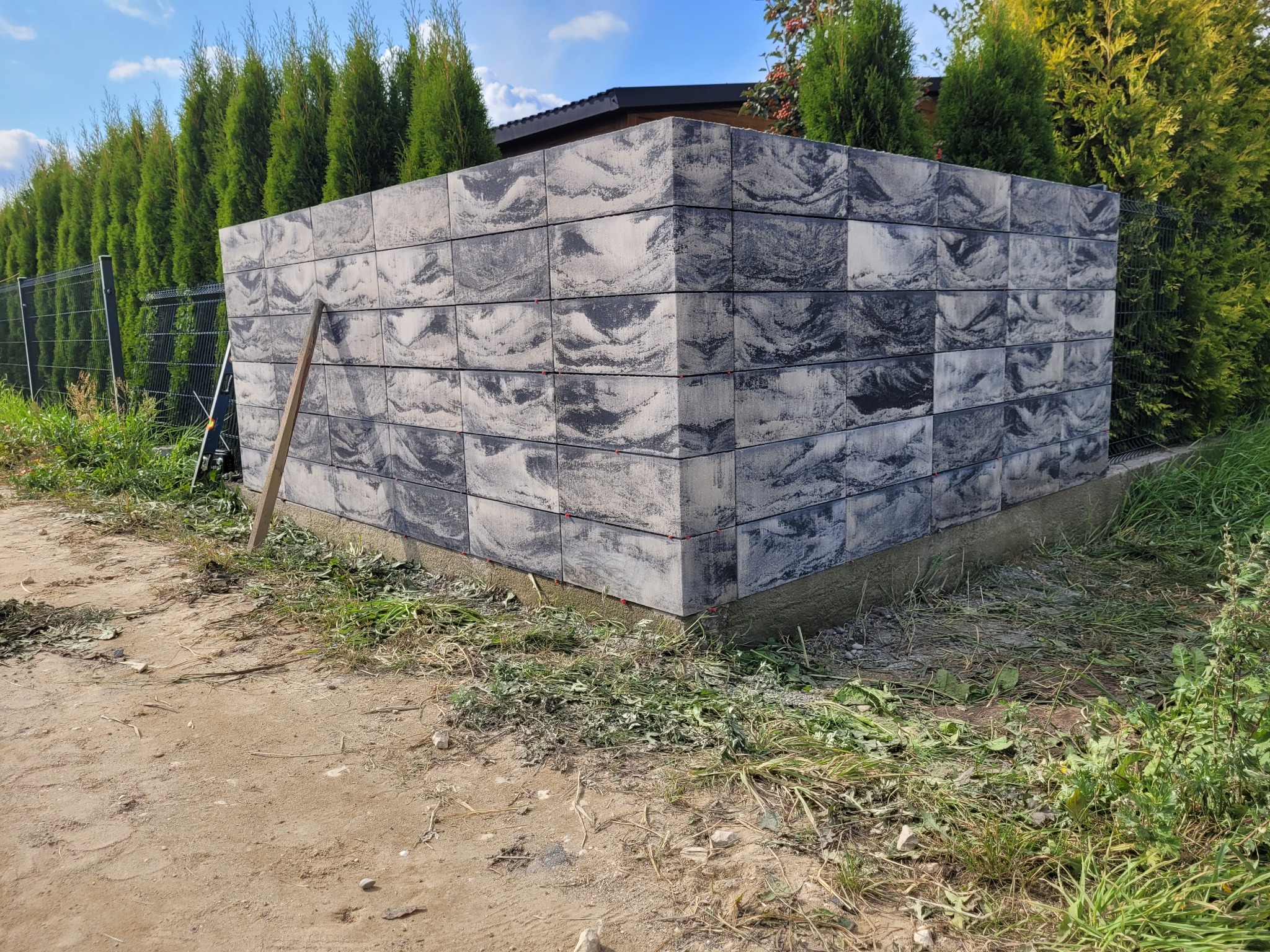 Wznoszony fragment ogrodzenia z szarych, wzorzystych bloczków betonowych, widoczne czerwone elementy poziomowania, w tle zielone tuje i metalowa siatka ogrodzeniowa.