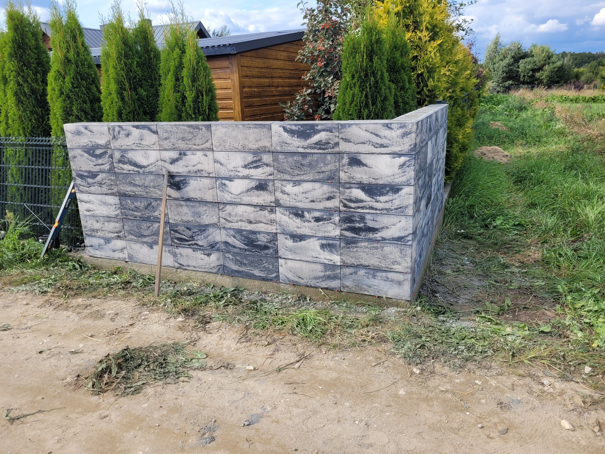 Fragment nowo postawionego, niskiego murku z szarych, wzorzystych bloczków betonowych na tle ogrodu z tujami i drewnianą altaną.