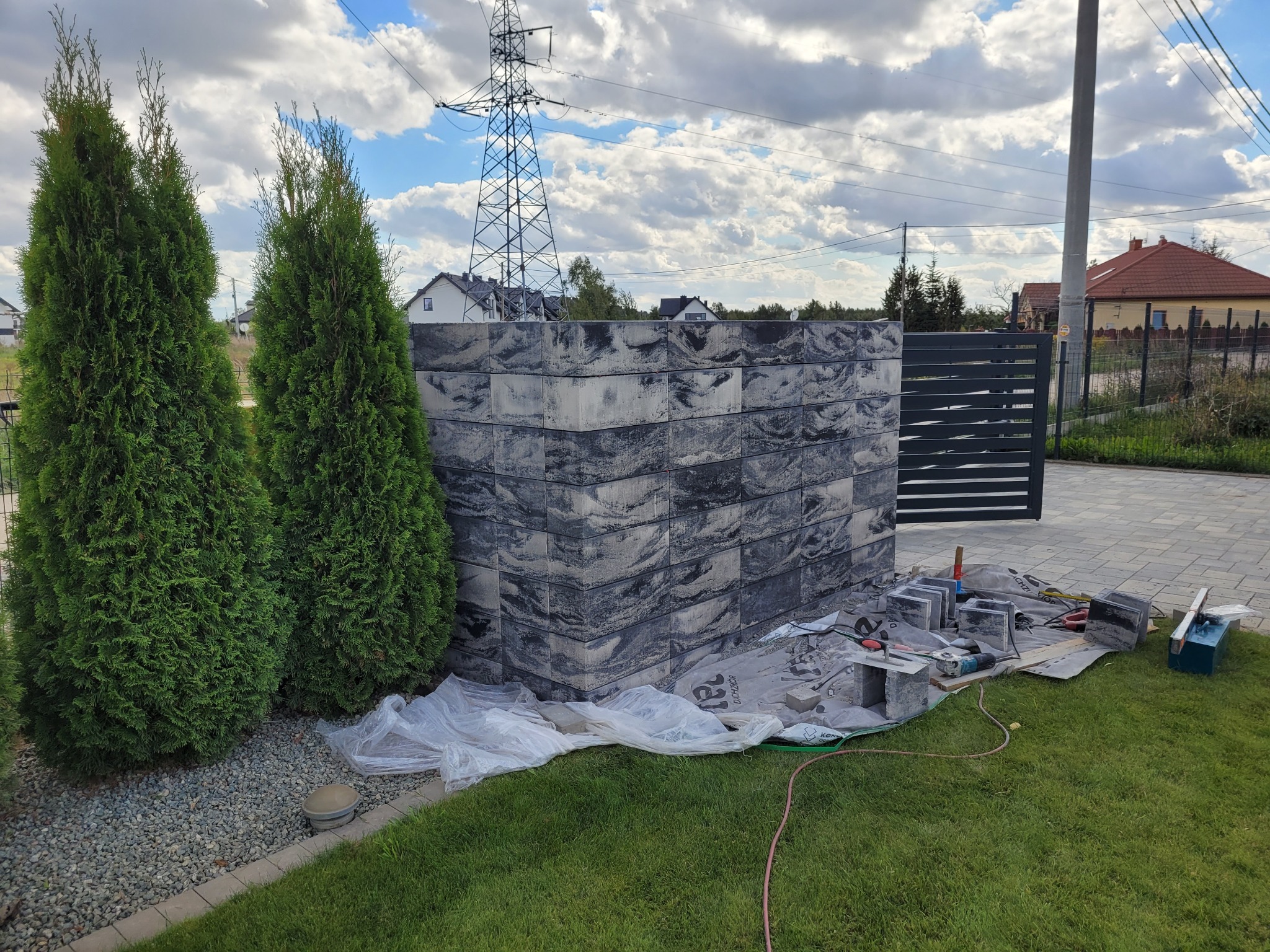 Budowa ogrodzenia z prefabrykatów betonowych o wzorze imitującym marmur, widoczne narzędzia i materiały budowlane, w tle słup wysokiego napięcia i domy.