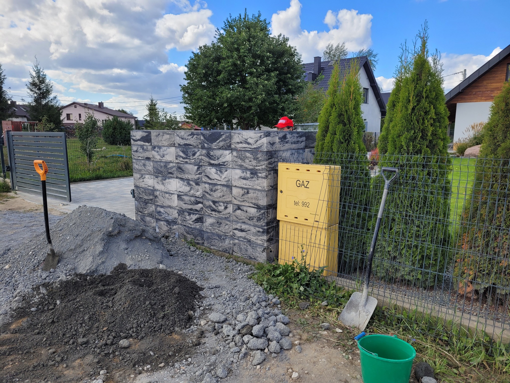 Wznoszony fragment ogrodzenia z szarej kostki łupanej, widoczna sterta ziemi, piasku, gruzu, dwie łopaty, wiadro, skrzynka gazowa na tle zieleni i domów.
