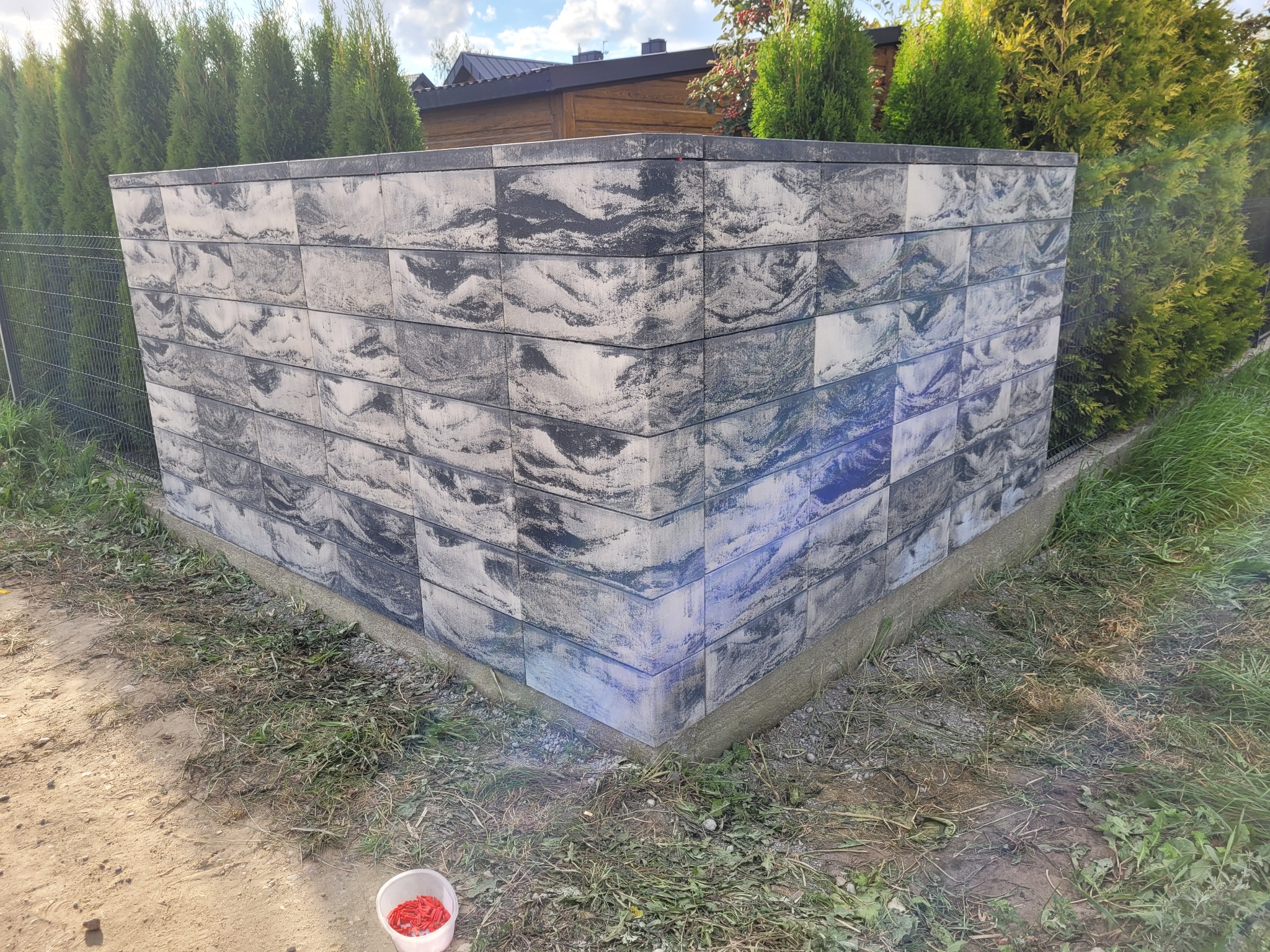 Fragment nowo wybudowanego ogrodzenia z betonowych bloczków o nieregularnym wzorze, widoczne tuje w tle, czerwone kołki w plastikowym wiaderku u podstawy.
