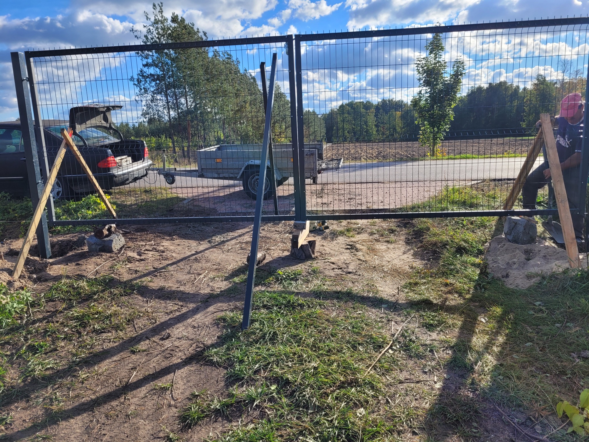 Proces instalacji bramy w ogrodzeniu panelowym: widoczne podparte słupki, auto z przyczepką w tle, pracownik przytrzymujący słupek.