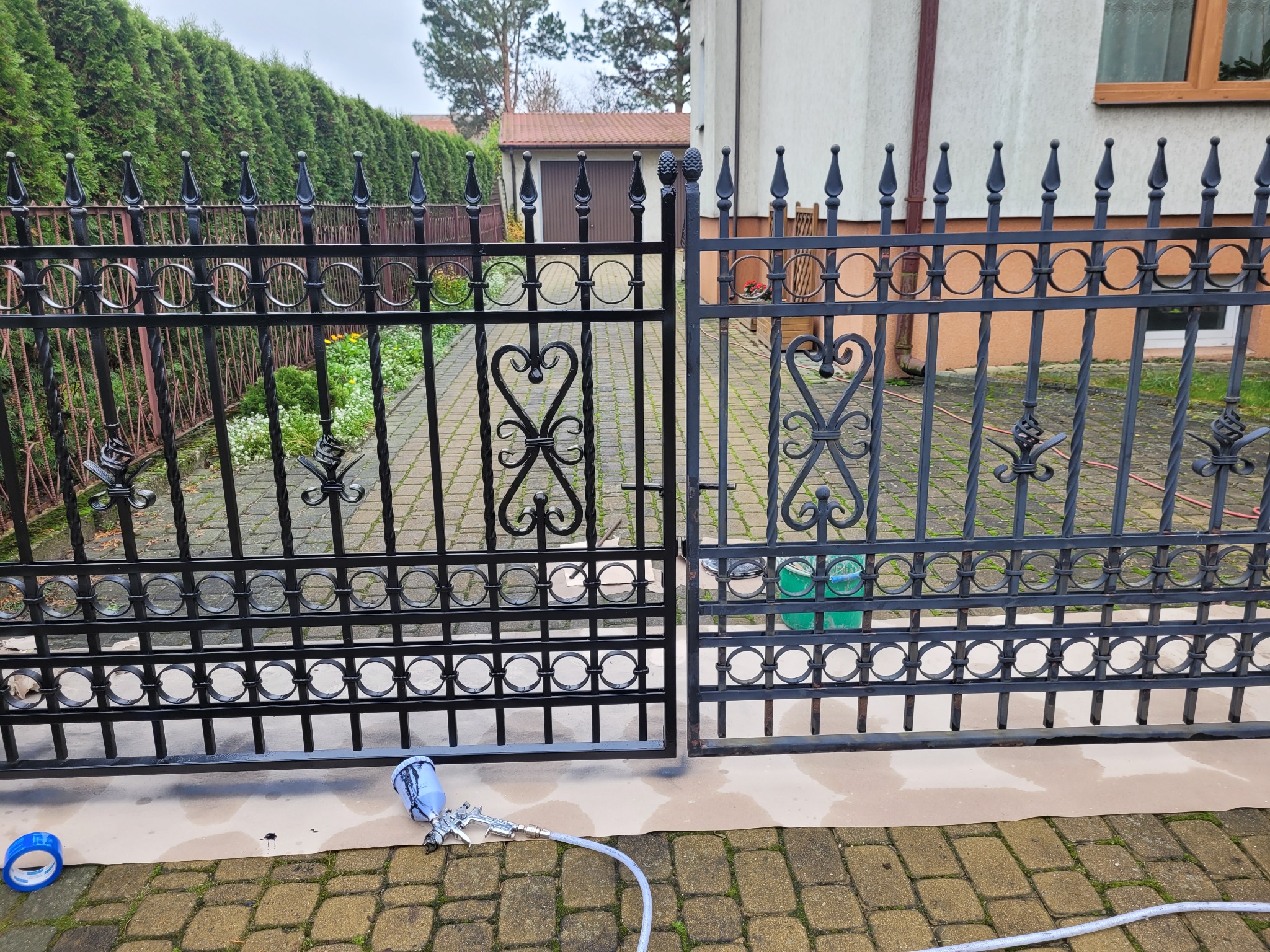 Malowanie natryskowe metalowej bramy wjazdowej na posesję, widoczny pistolet lakierniczy, zabezpieczone podłoże, częściowo odnowiona konstrukcja.
