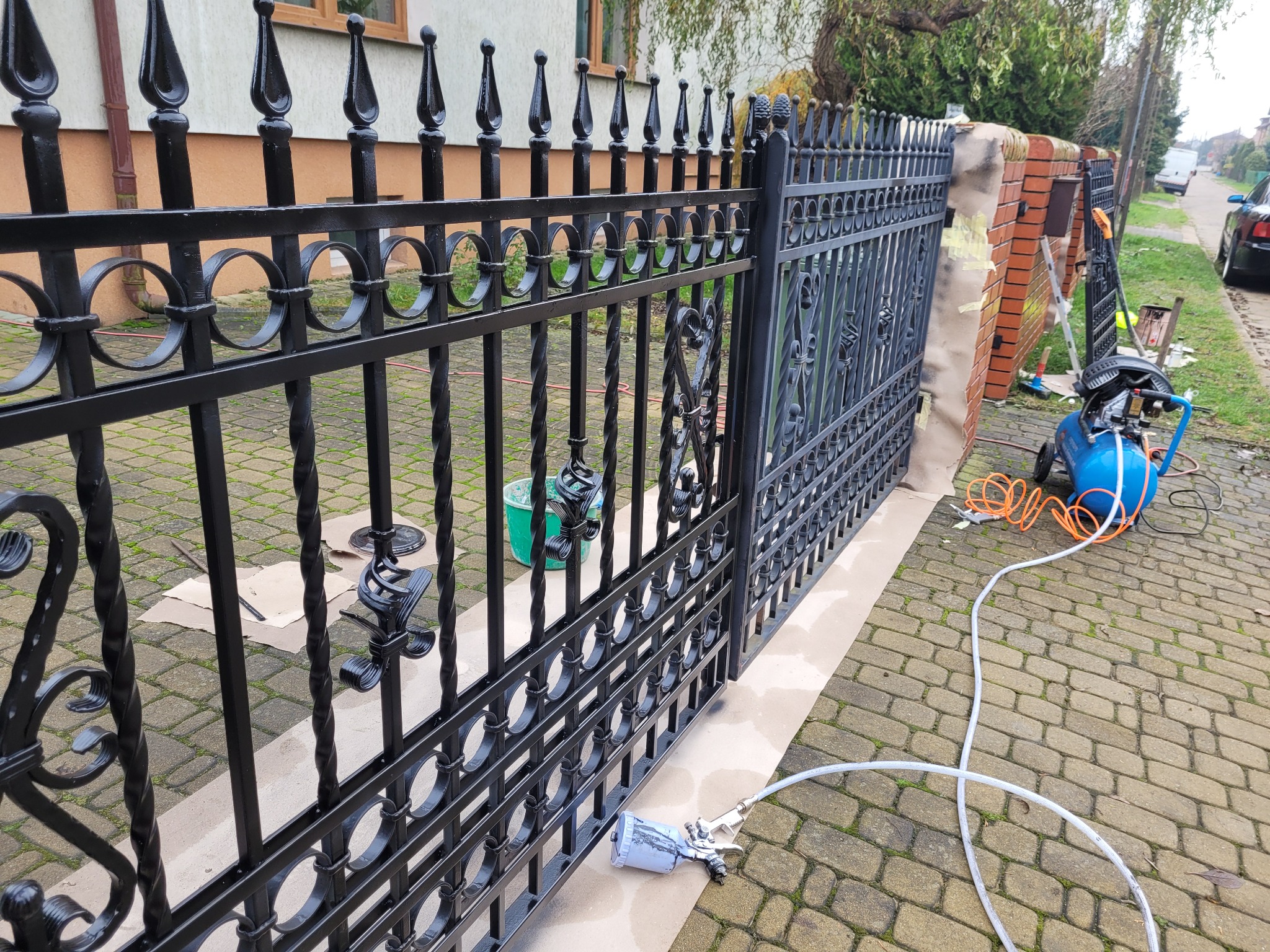 Czarny, metalowy płot z ozdobnymi elementami w trakcie malowania natryskowego; widoczny kompresor, pistolet lakierniczy i wiadro z farbą.