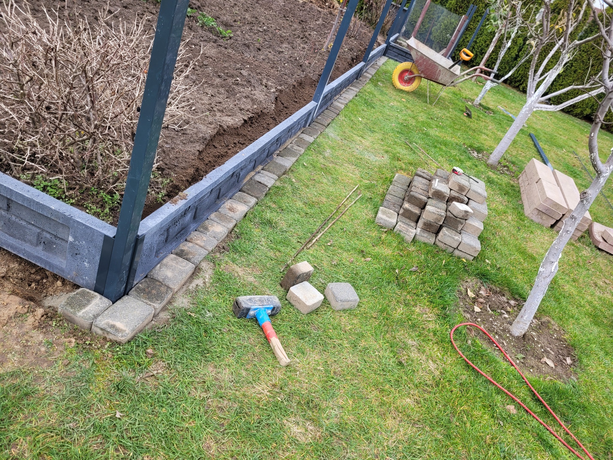 Budowa ogrodzenia z betonowych elementów na trawniku, widoczny fragment konstrukcji ogrodzenia, paleta kostek, gumowy młotek i taczka w tle.