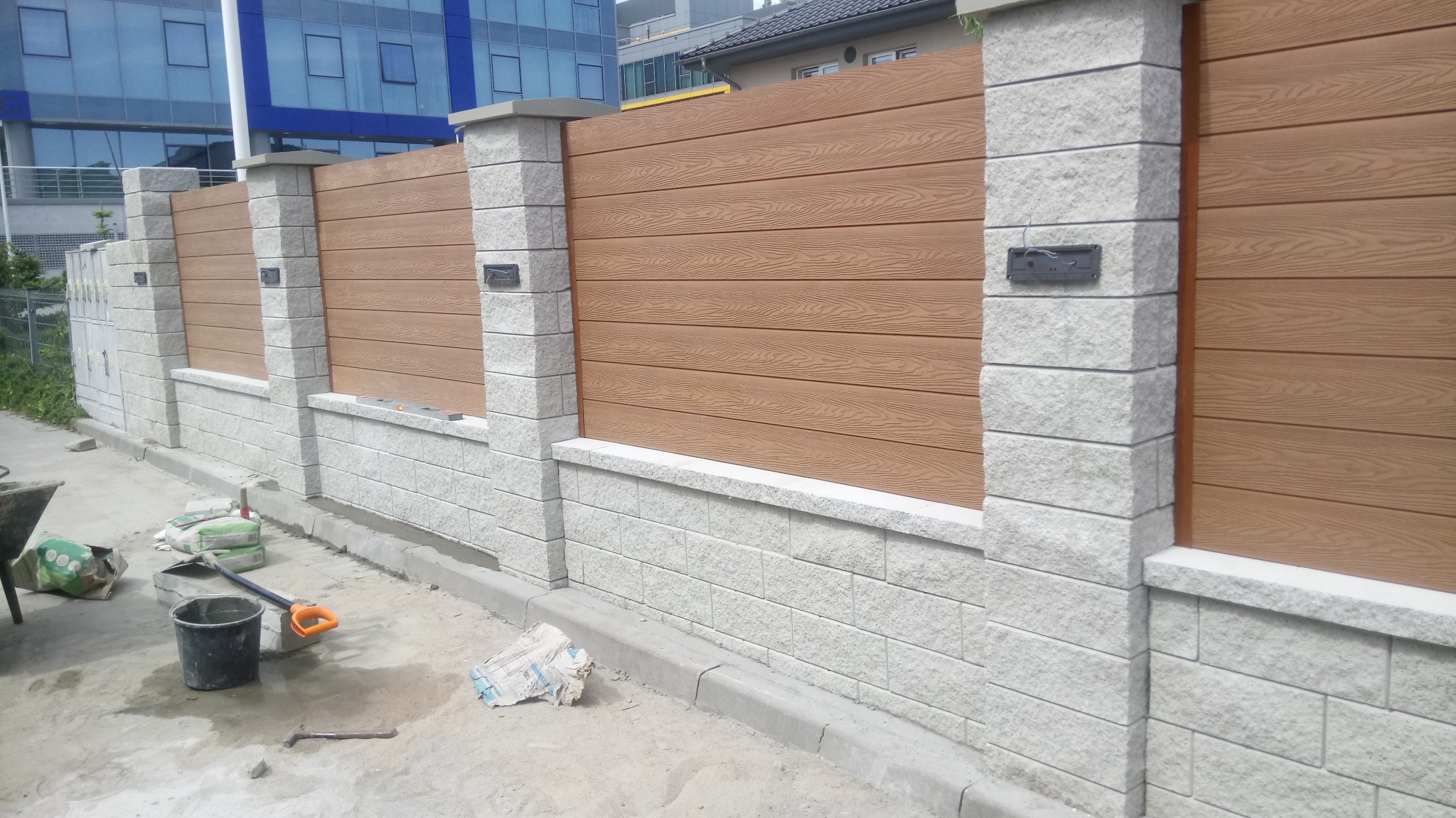 Montaż ogrodzenia z paneli imitujących drewno i kamiennych słupków, widoczne narzędzia pracy i worki z materiałami.