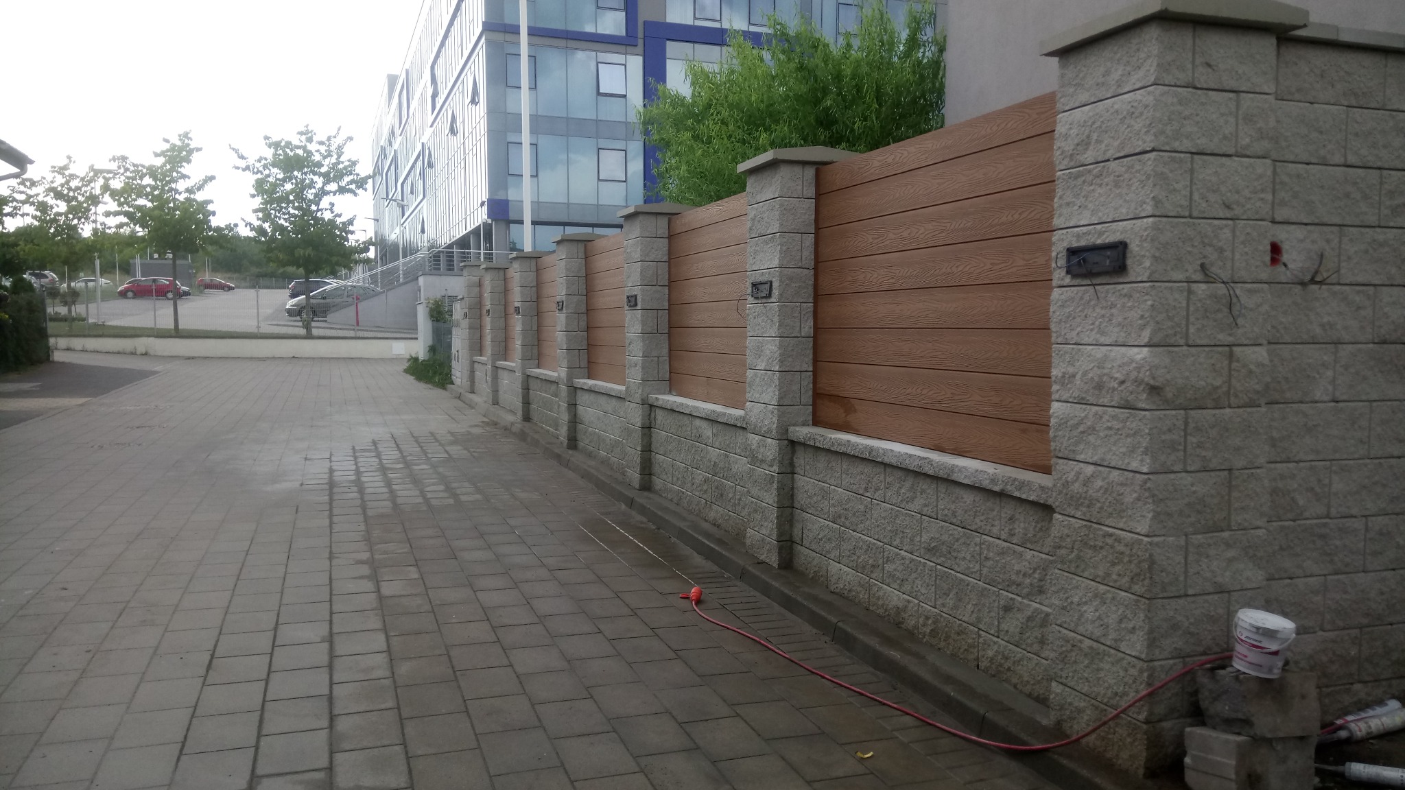 Nowoczesne ogrodzenie z paneli imitujących drewno i kamiennych słupków, z widocznym przewodem i pojemnikiem po farbie przy podstawie, na tle biurowca.