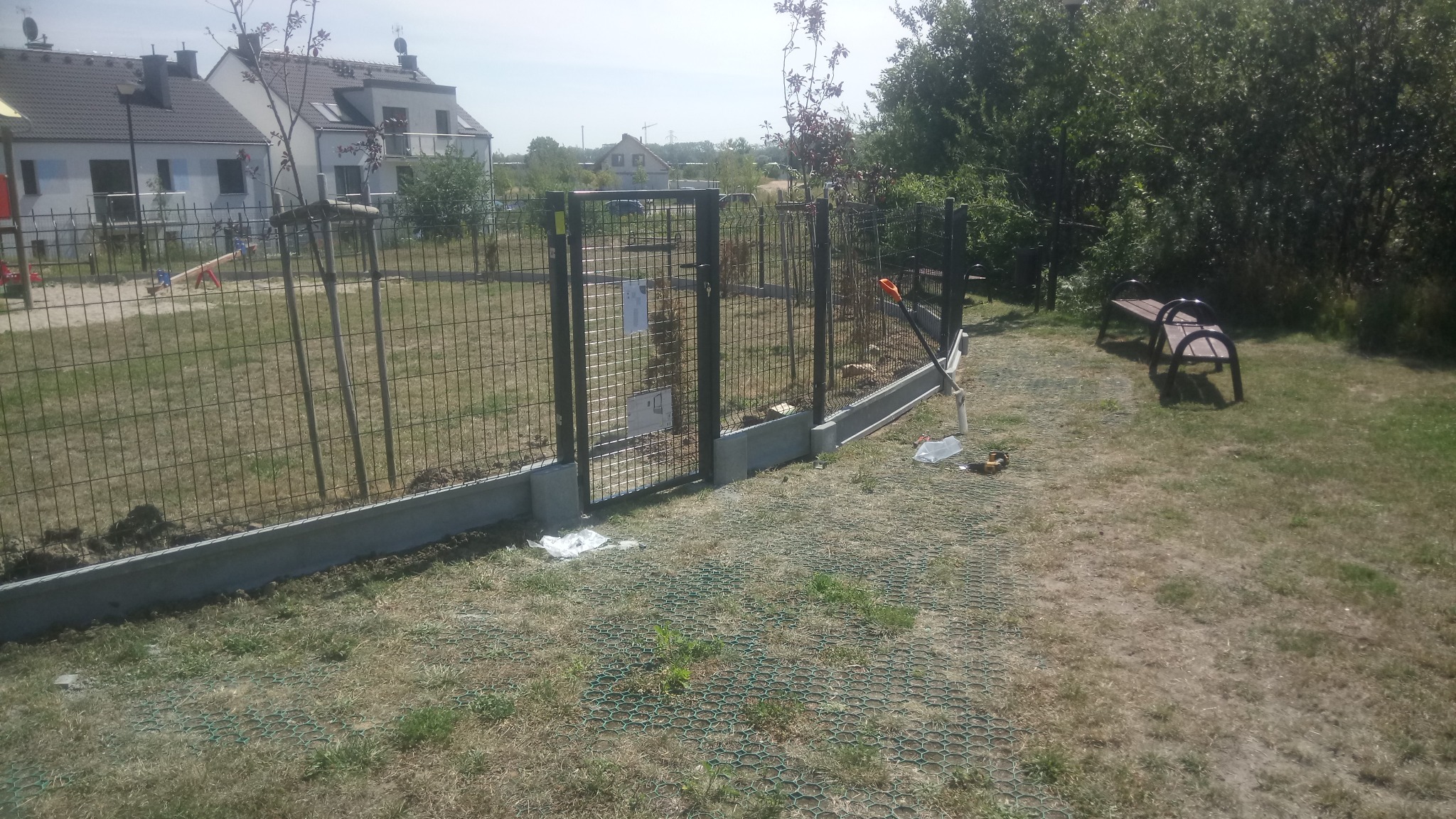 Nowo postawione ogrodzenie z siatki z betonową podmurówką i furtką, widoczne narzędzia i resztki materiałów po montażu, w tle domy mieszkalne i drzewa.