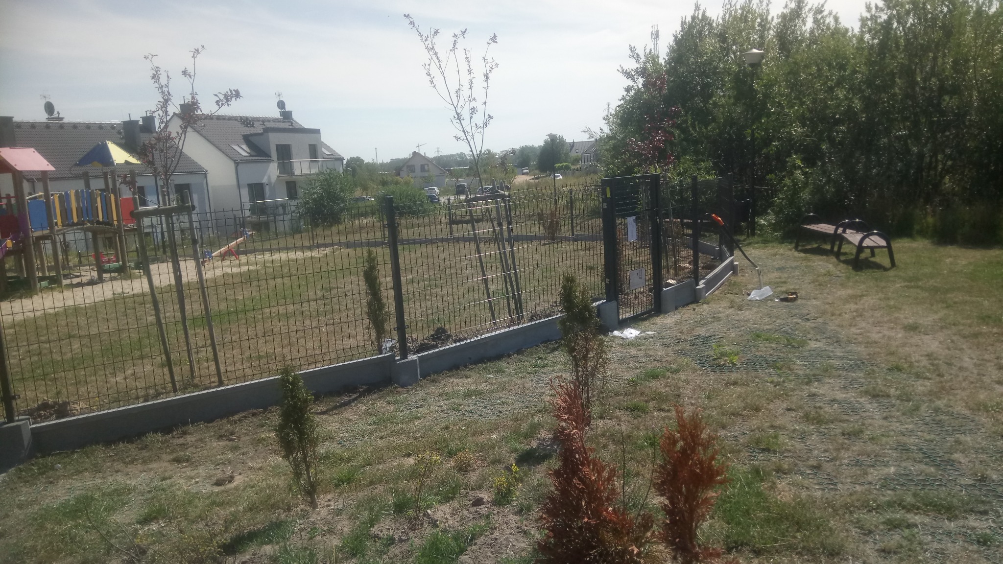 Nowo postawione metalowe ogrodzenie panelowe z podmurówką betonową na terenie posesji z placem zabaw i ławką, widoczne narzędzia ogrodnicze.