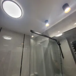 Nowoczesna łazienka z szarą marmurową okładziną, kabiną prysznicową ze szkła z czarnymi ramami i oświetleniem sufitowym: okrągły panel LED i dwa reflektory punktowe.