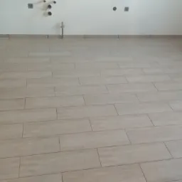 Ułożona podłoga z podłużnych, beżowych płytek imitujących drewno, widoczne przyłącza wodne i elektryczne na białej ścianie w tle.