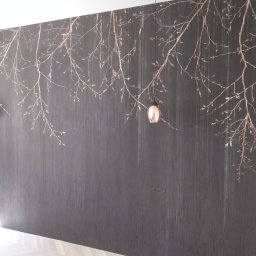 Roman&Ko - Ciemnoszara ściana z pionowymi paskami i motywem gałęzi z pąkami w kolorze miedzi, dwie wiszące lampy z miedzianymi kloszami, jasna podłoga z parkietu.