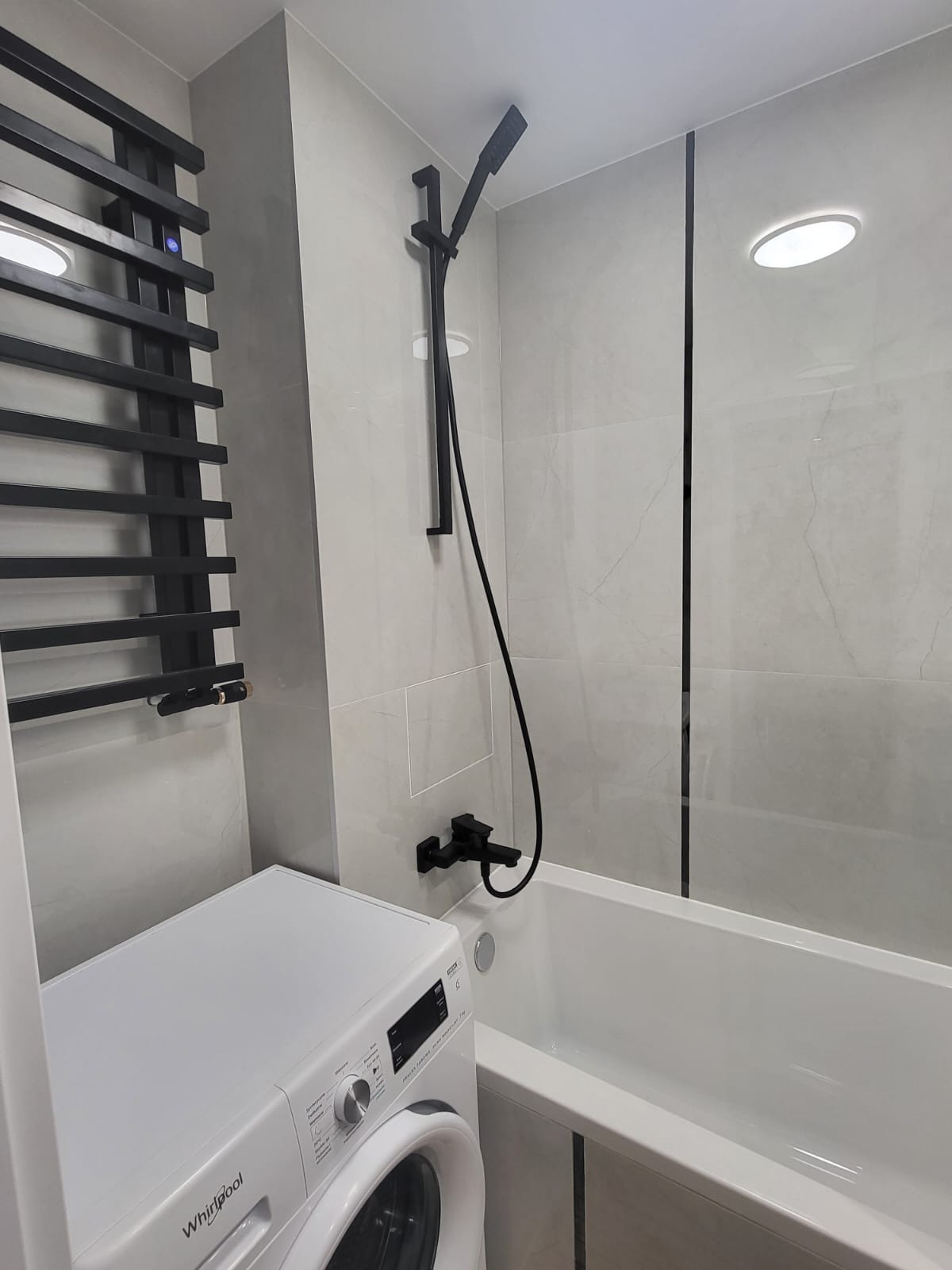 Nowoczesna łazienka z pralką Whirlpool, czarną armaturą, grzejnikiem drabinkowym i wanną obudowaną płytkami imitującymi marmur.