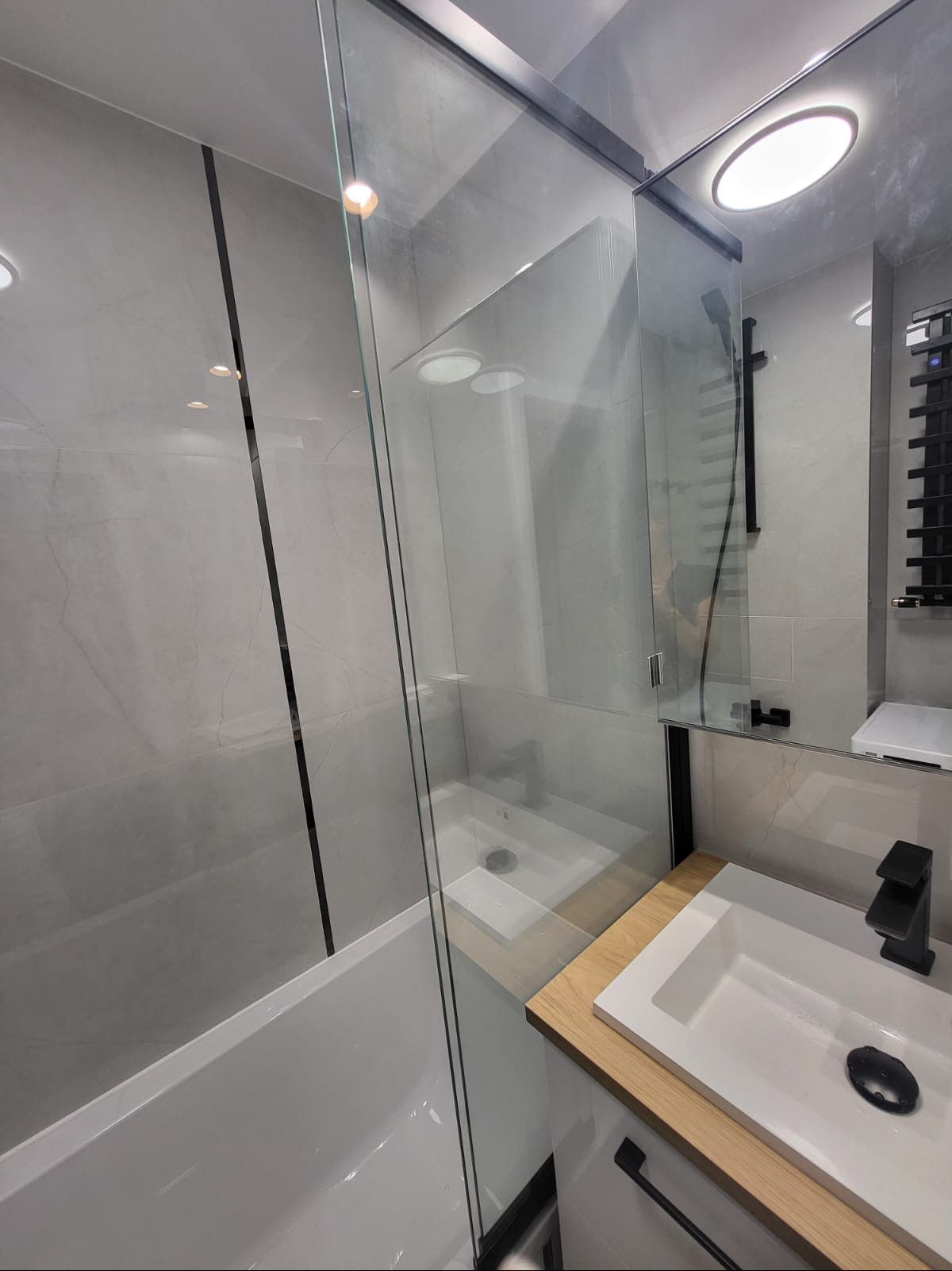 Nowoczesna łazienka z kabiną prysznicową typu walk-in, szare płytki imitujące marmur, czarna armatura i grzejnik drabinkowy, oświetlenie punktowe.