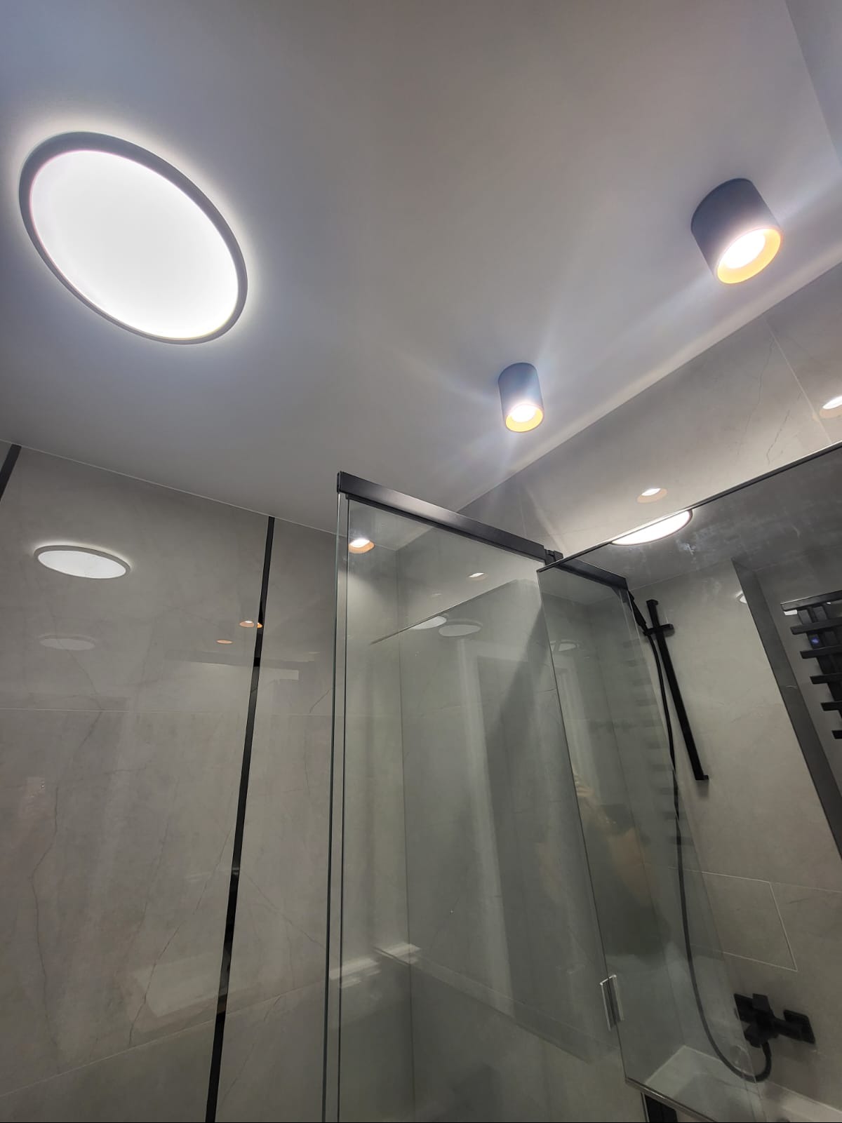 Nowoczesna łazienka z szarą marmurową okładziną, kabiną prysznicową ze szkła z czarnymi ramami i oświetleniem sufitowym: okrągły panel LED i dwa reflektory punktowe.