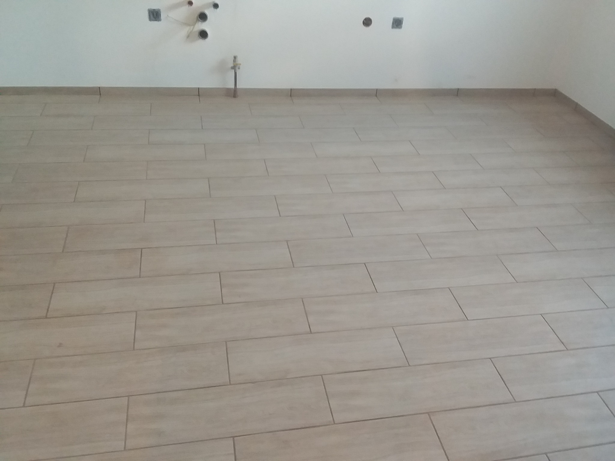 Ułożona podłoga z podłużnych, beżowych płytek imitujących drewno, widoczne przyłącza wodne i elektryczne na białej ścianie w tle.