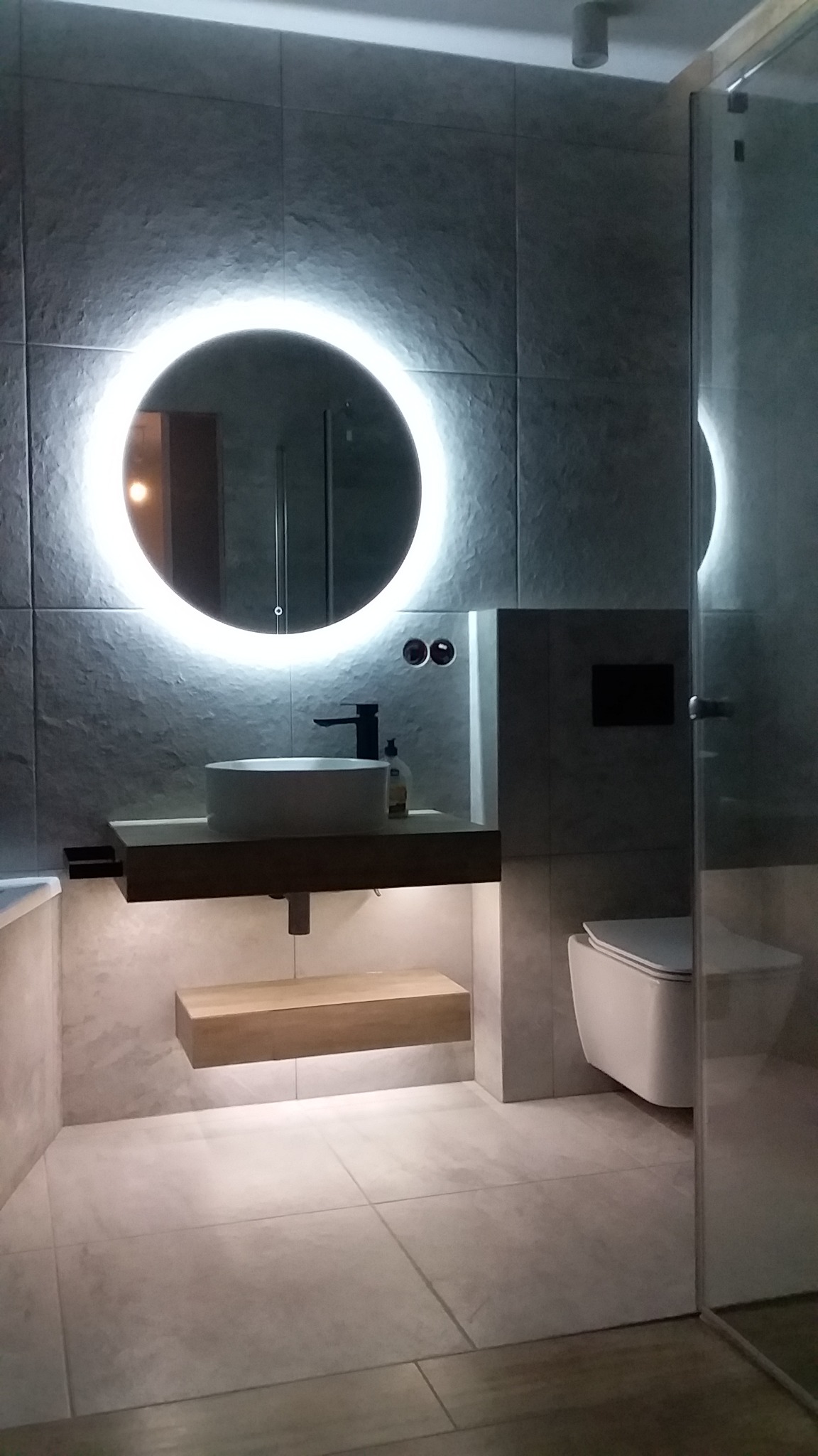 Nowoczesna łazienka z okrągłym lustrem podświetlanym LED, wiszącą umywalką na drewnianym blacie, toaletą podwieszaną i szarymi płytkami imitującymi kamień na ścianach.