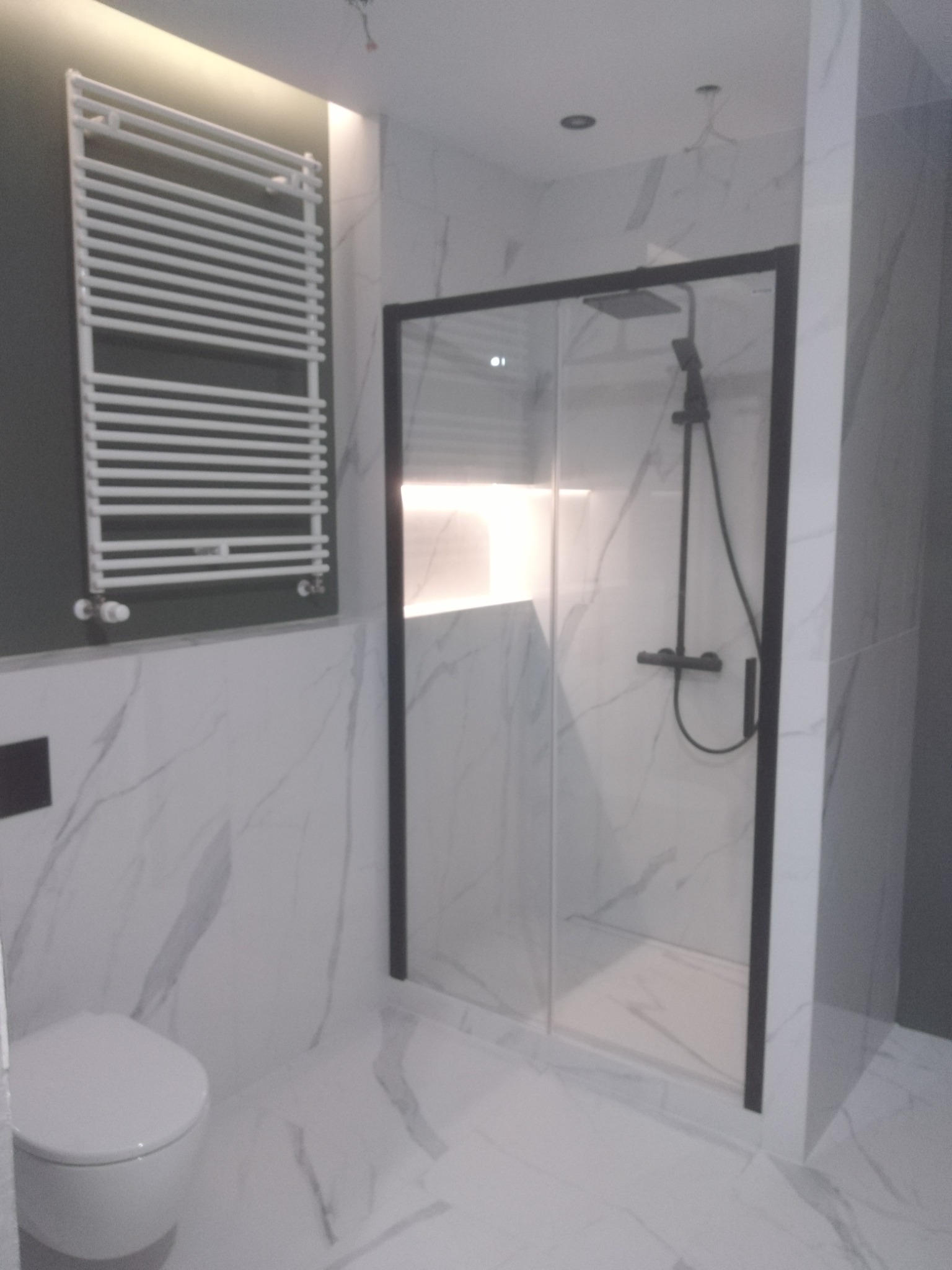 Nowoczesna łazienka z kabiną prysznicową w czarnej ramie, grzejnikiem drabinkowym na szarej ścianie i toaletą wiszącą, wykończona białymi płytkami imitującymi marmur.