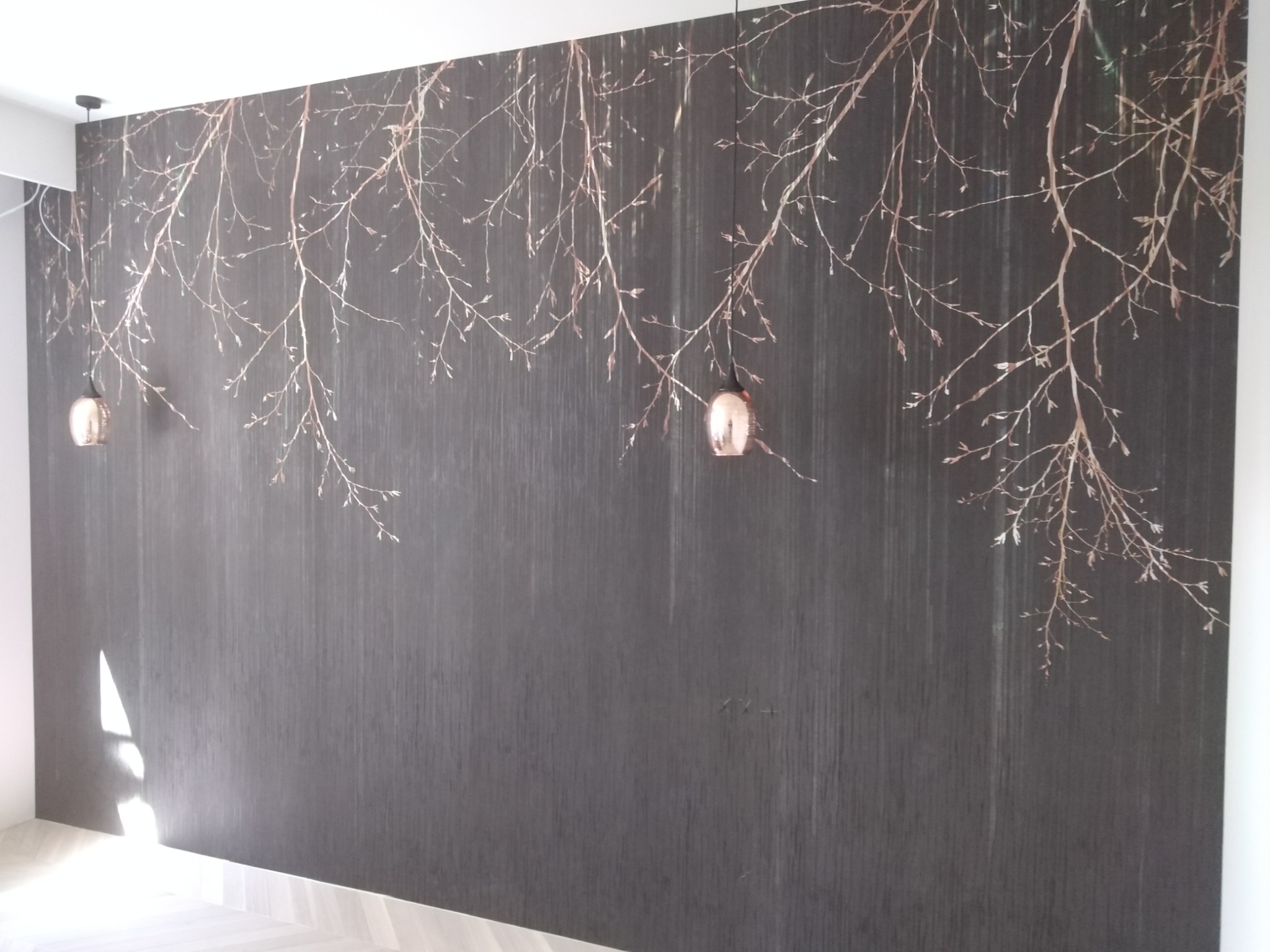 Ciemnoszara ściana z pionowymi paskami i motywem gałęzi z pąkami w kolorze miedzi, dwie wiszące lampy z miedzianymi kloszami, jasna podłoga z parkietu.
