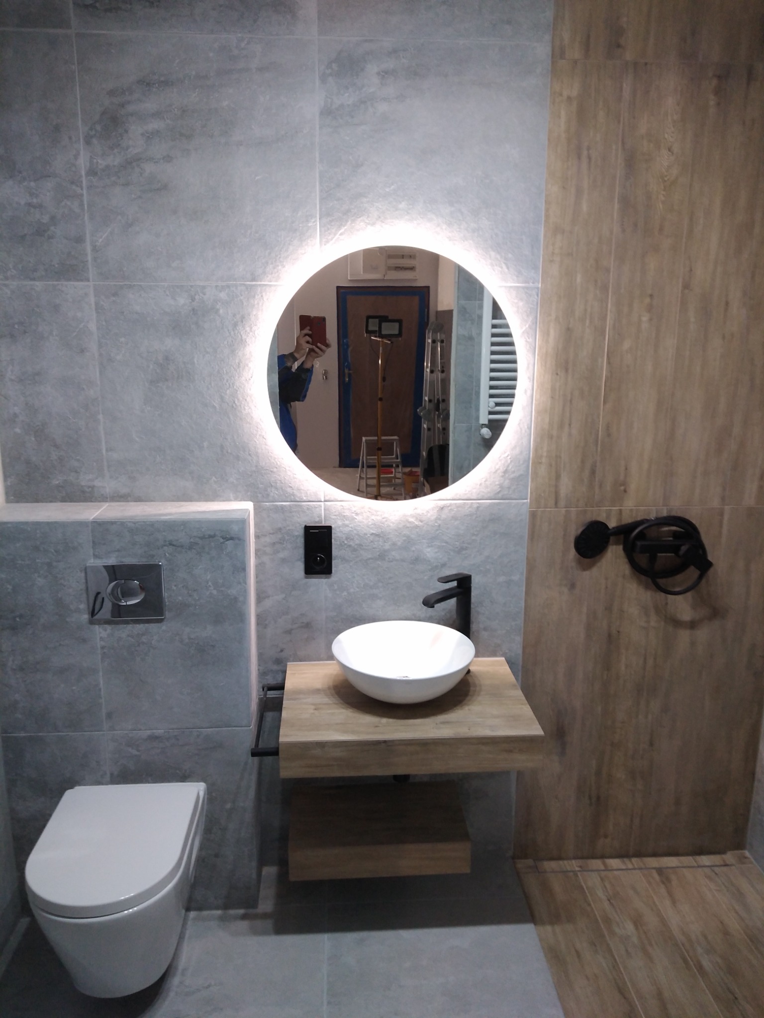 Nowoczesna łazienka z szarymi płytkami imitującymi kamień, okrągłym podświetlanym lustrem, wiszącą białą umywalką na drewnianym blacie i podwieszaną toaletą, odzwierciedlenie fotografa w lustrze.